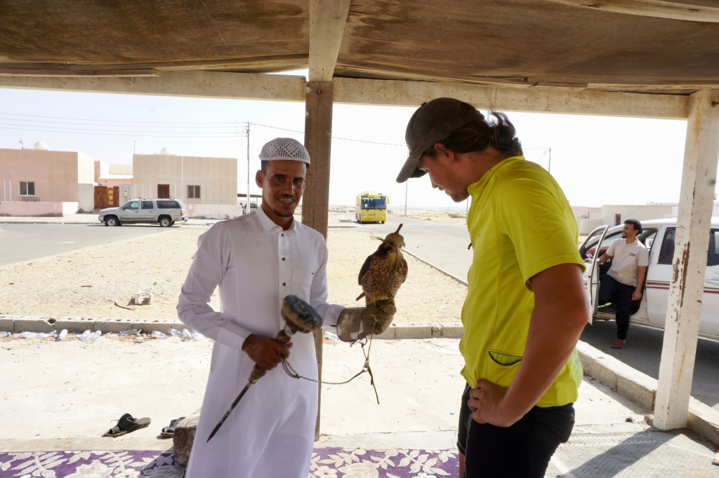 A man in a white Saudi Arabian thobe standing next to another man. The man on the left has a live falcon sitting on his forearm.